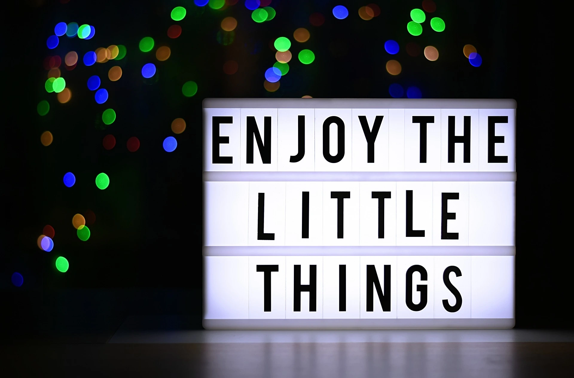 Ein Leuchtschild mit der Aufschrift: Enjoy the little things. Dahinter ein schwarzer Hintergrund mit bunten Punkten.
