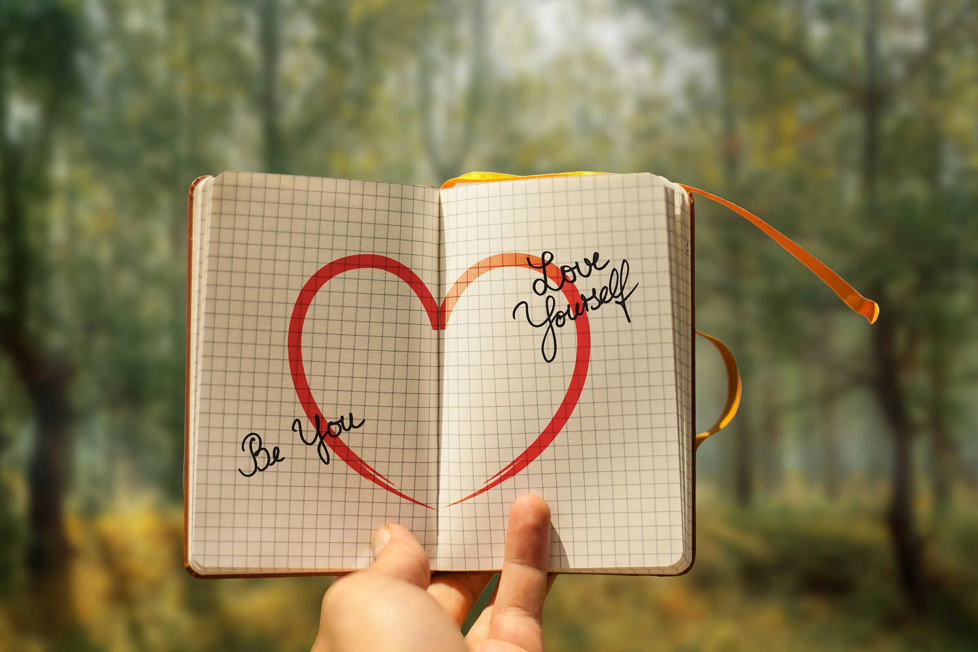 Ein Notizbuch in das über beide Seiten ein Herz gemalt wurde. Links steht 'Be you', rechts 'Love yourself'.